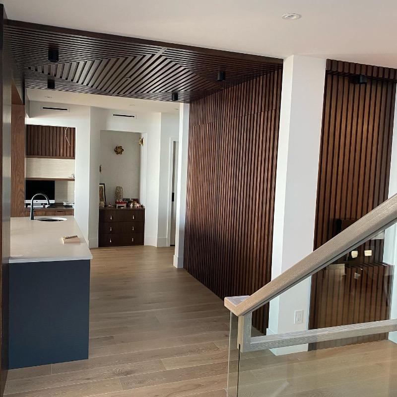 Walnut slat panelling running across a wall and ceiling beside an open-plan kitchen and glass-railed staircase